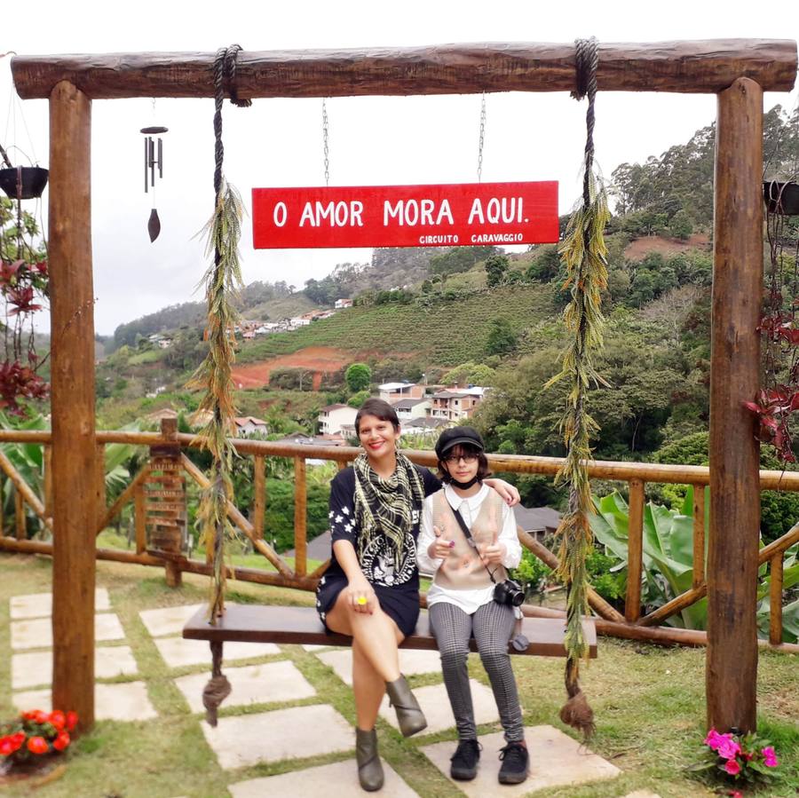 Milla Camata e a filha sentadas num balanço de madeira sob uma placa vermelha escrita O amor mora aqui - Circuito Caravaggio, com o vale ao fundo