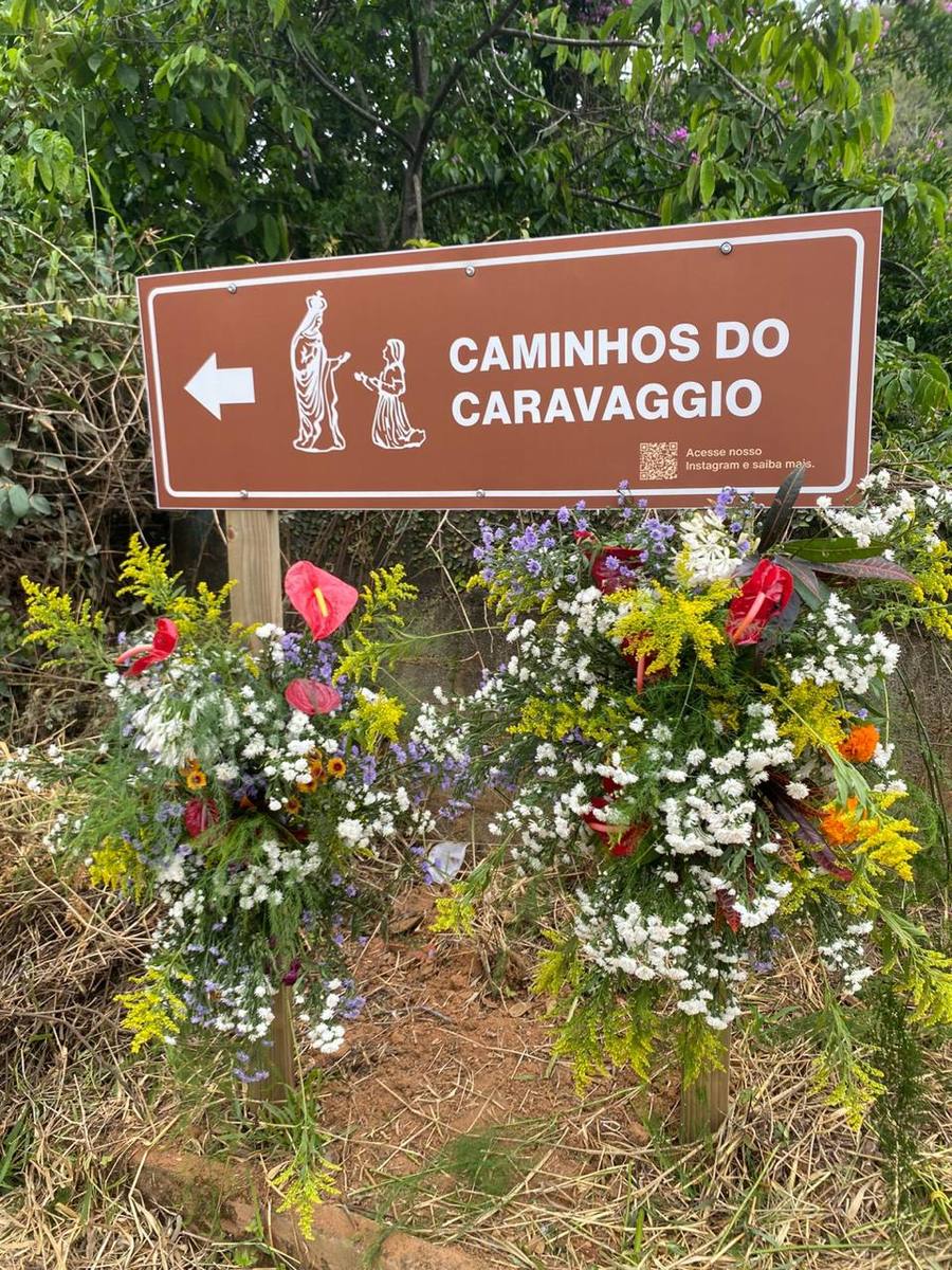 Placa marrom de sinalização turística com os dizeres Caminhos do Caravaggio, cercada de arranjos de flores na beira da estrada