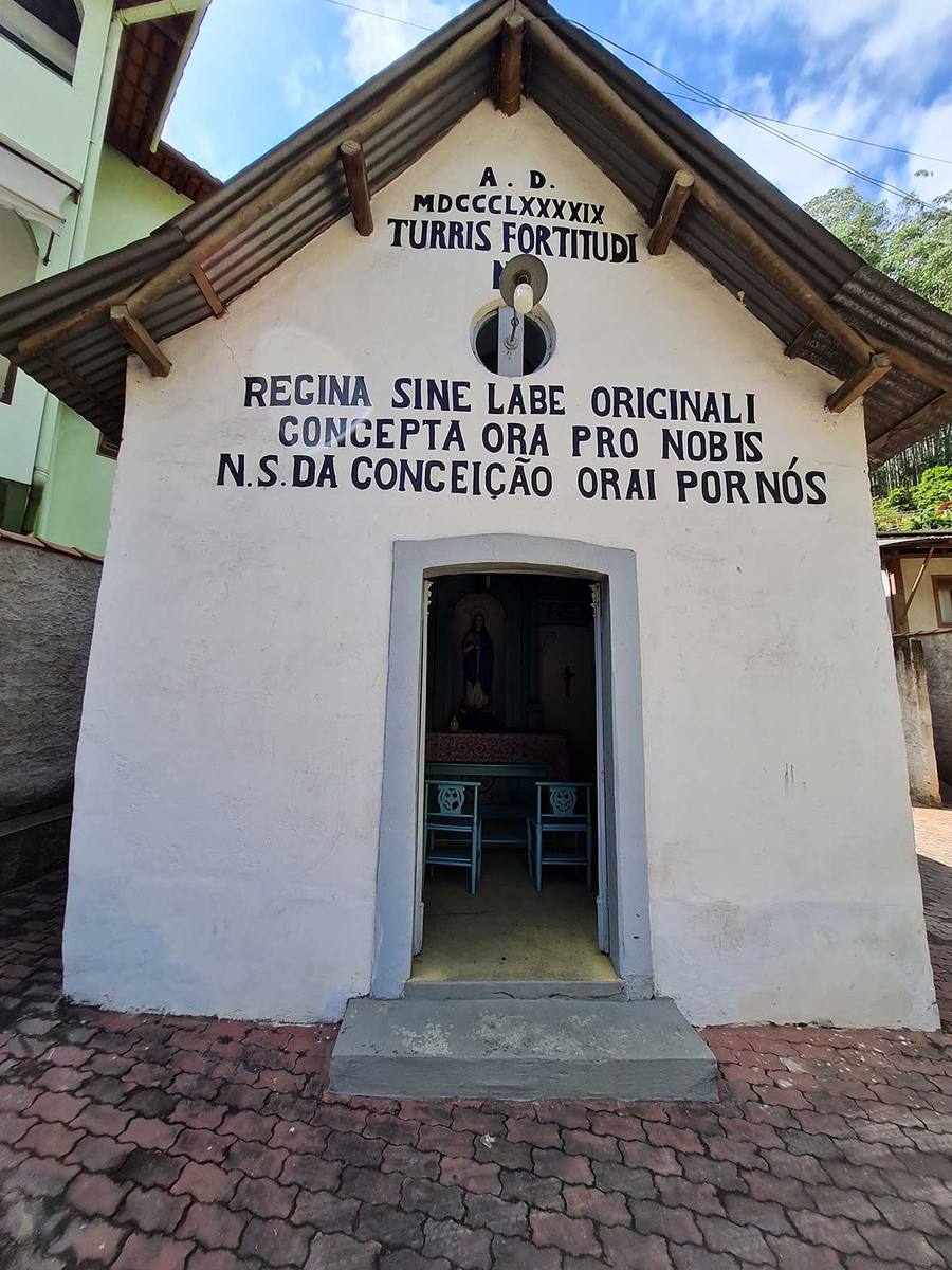 Fachada branca da Capela de Nossa Senhora da Conceição, de 1899, com inscrições em latim e português e a imagem da santa visível pela porta aberta