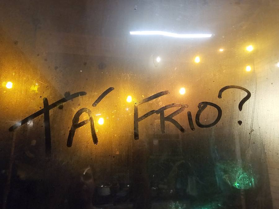 A pergunta Tá frio? escrita com o dedo no vidro embaçado de uma janela, com luzes amarelas ao fundo