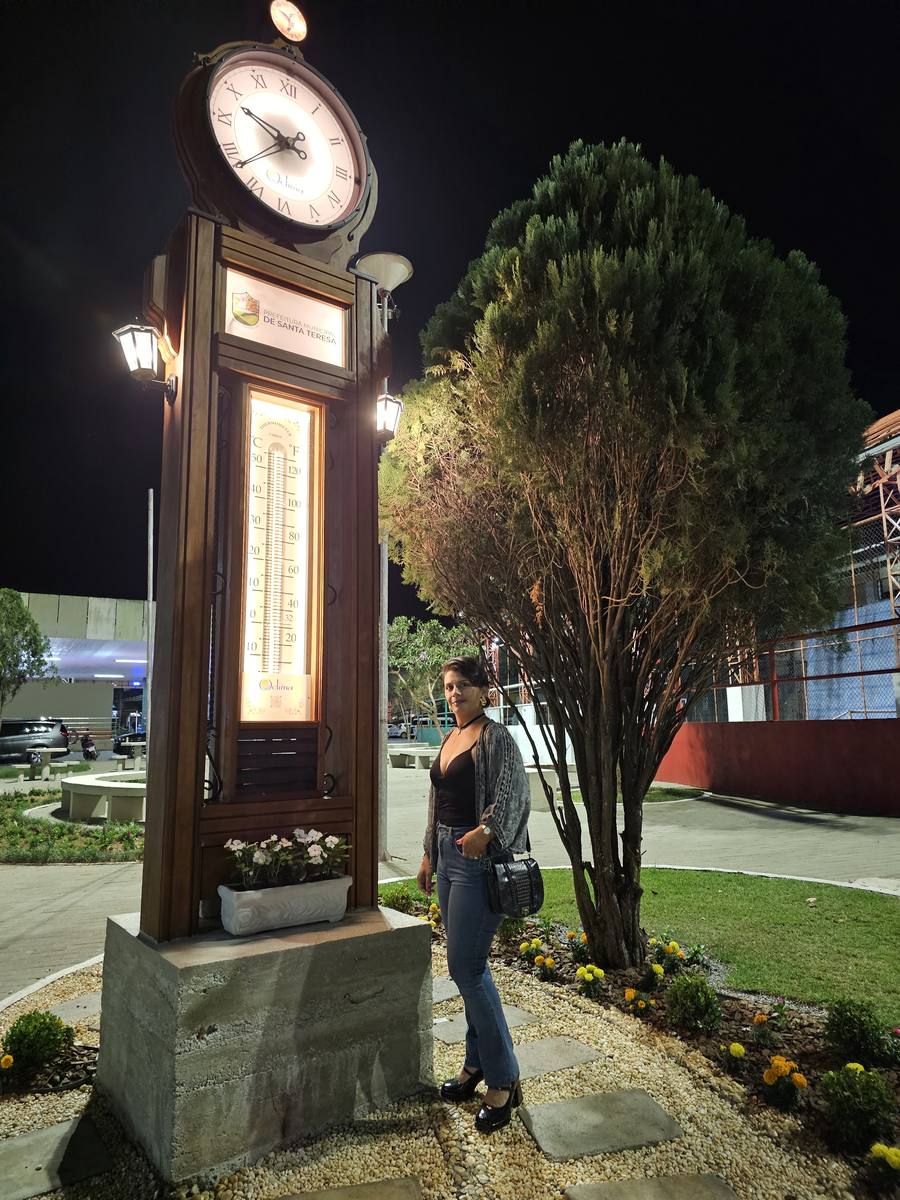 Milla Camata ao lado do relógio com termômetro da Prefeitura de Santa Teresa, iluminado à noite na praça