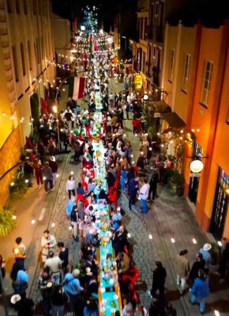 Vista do alto da Rua do Lazer à noite, com uma mesa única atravessando a rua inteira, iluminação suspensa e bandeiras italianas