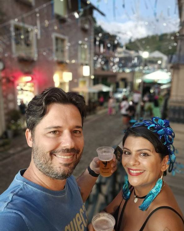 Casal brindando com chope na calçada da Rua do Lazer durante o Carnaval, com fantasias e decoração de rua ao fundo