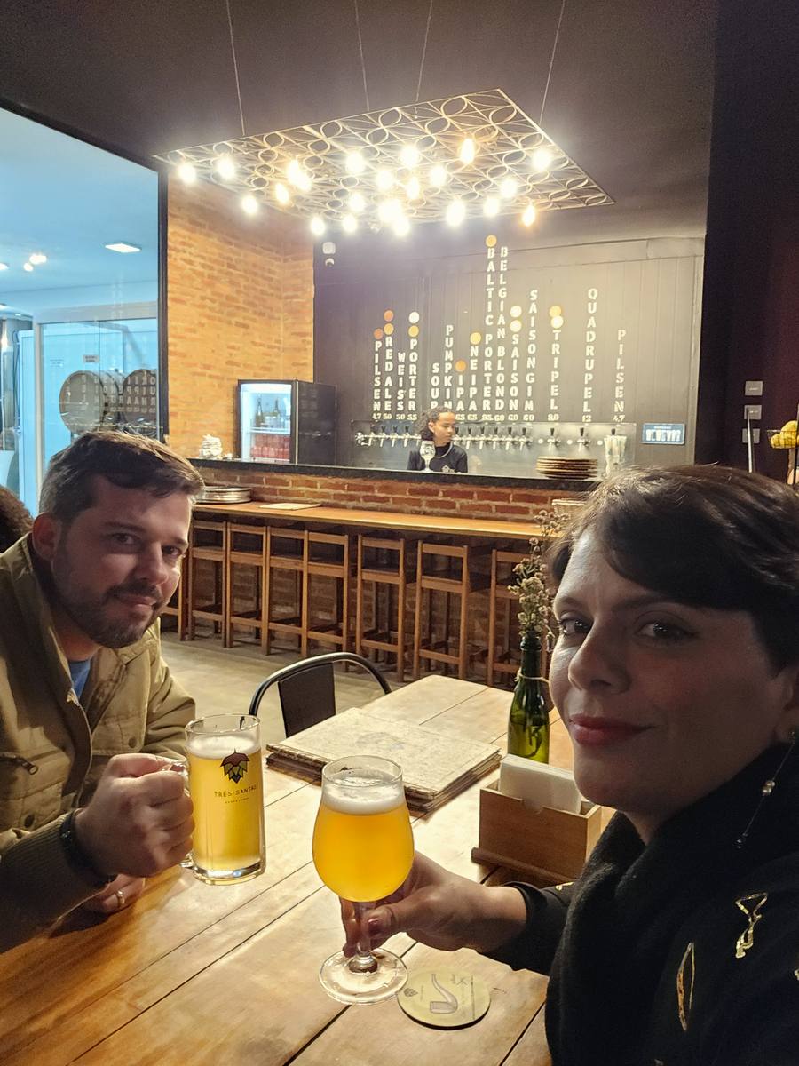 Mesa com duas cervejas servidas e, ao fundo, o painel com os estilos disponíveis na torneira do balcão da Três Santas