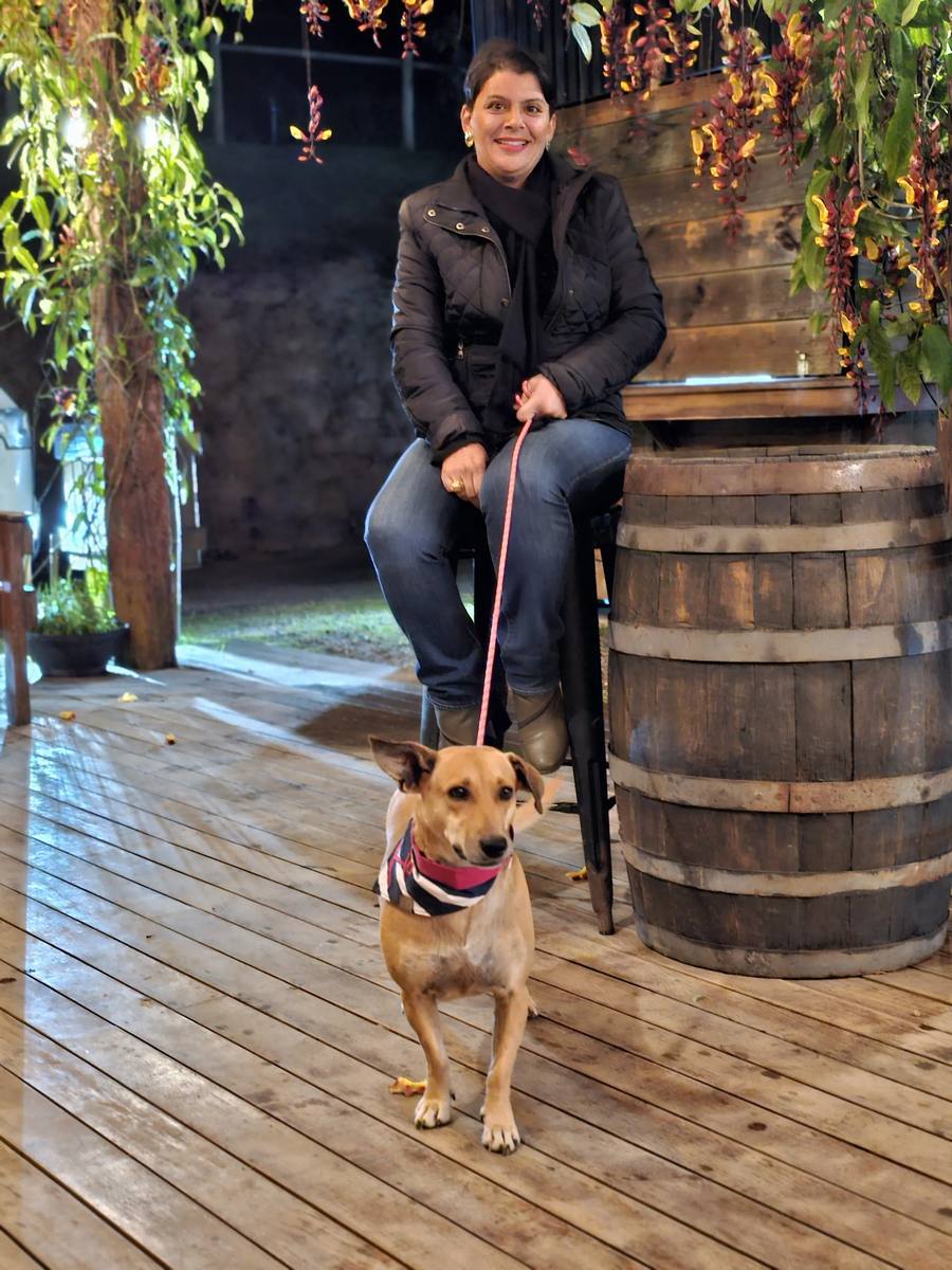Milla Camata sentada em um banco alto ao lado de um barril de madeira, com a cachorra na guia, no deck da cervejaria à noite
