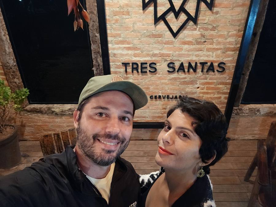 Leonardo e Milla diante do letreiro Três Santas Cervejaria, em parede de tijolo aparente