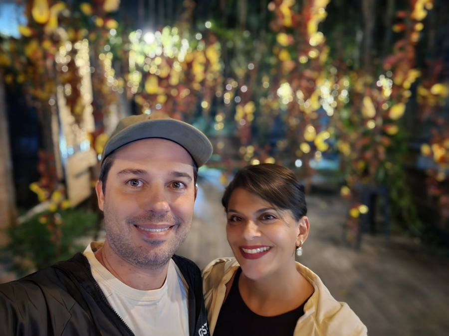 Leonardo e Milla sorrindo sob trepadeiras floridas iluminadas por luzes amarelas, na área externa da cervejaria